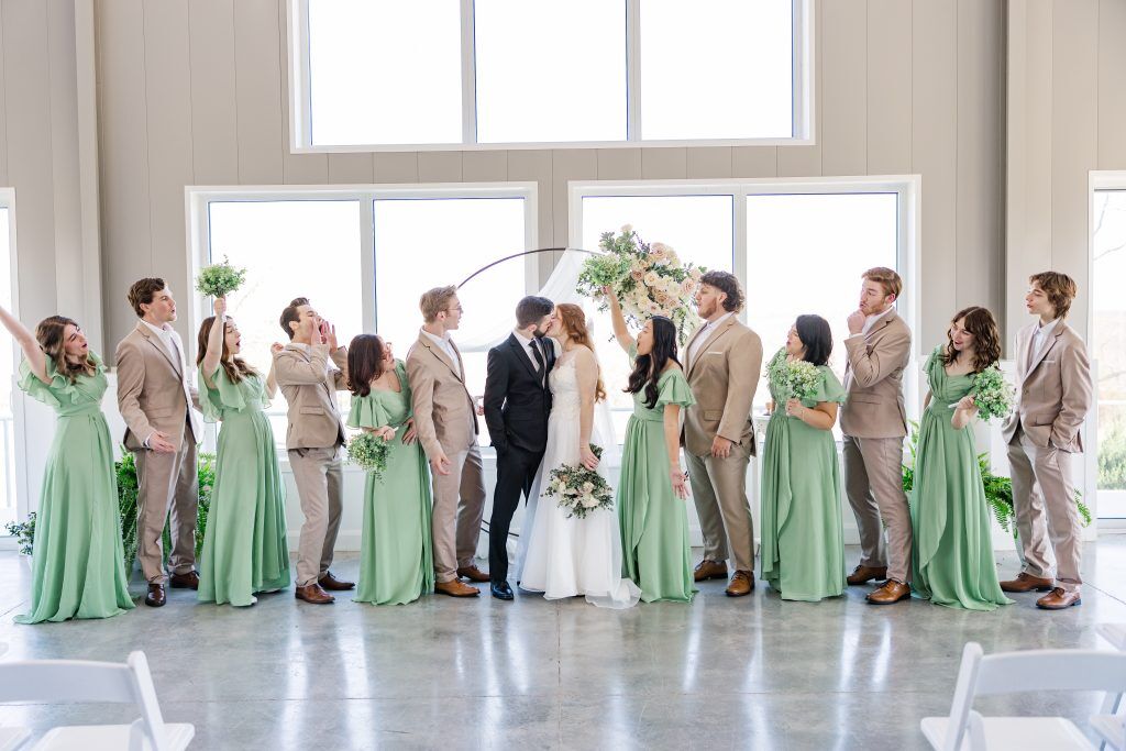 Beautiful wedding party of 14 dressed in Green and Light beige tuxedos in front of big windows at Bella Rosa Venue near Tulsa OK